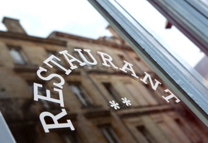 Close-up of a glass restaurant window with the word “RESTAURANT” and two asterisks in white lettering, reflecting a historic stone building.